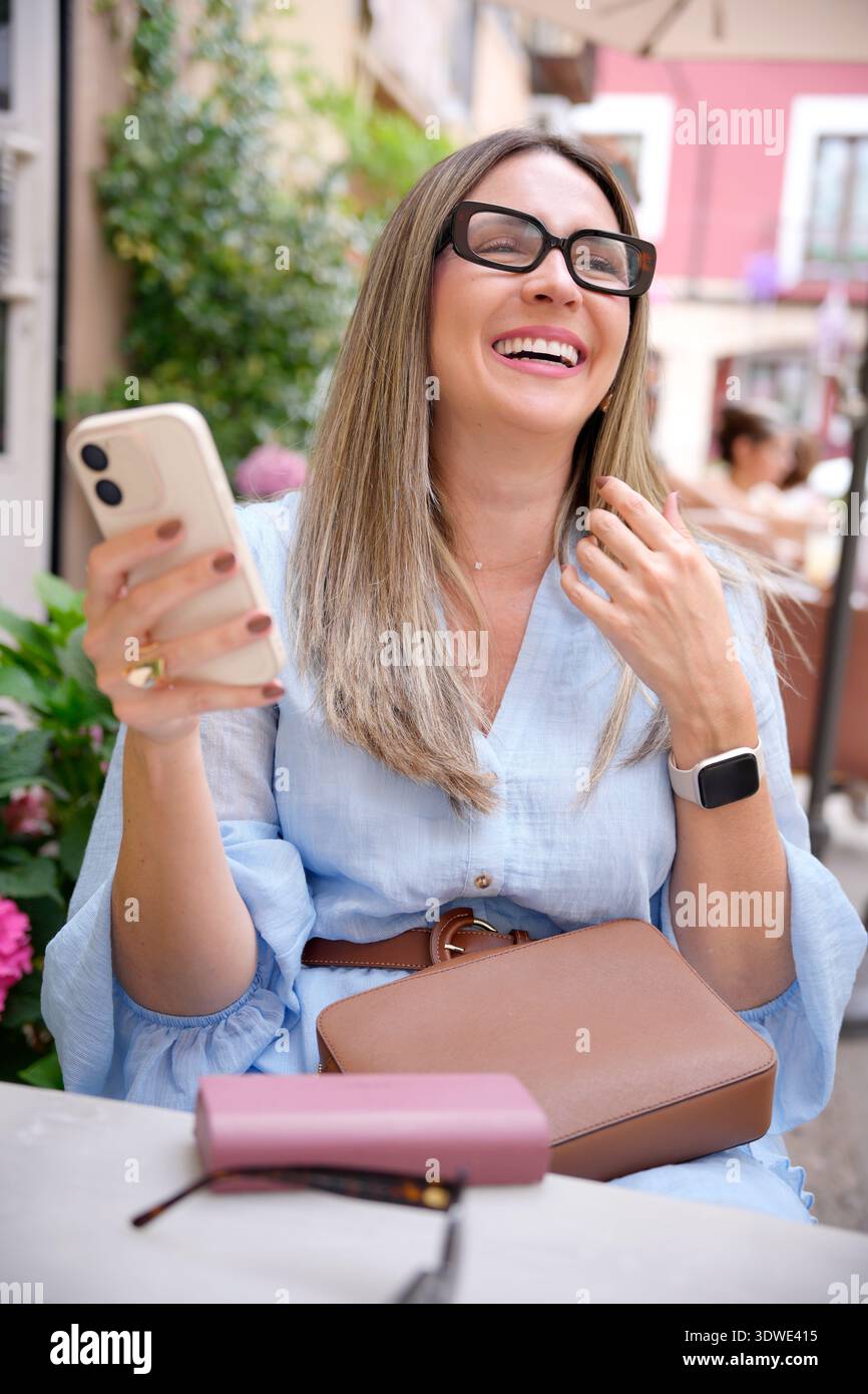 Une femme mature dans un café profitant d'une journée ensoleillée, souriante et tenant un smartphone. Elle porte des lunettes et une smartwatch, créant un a joyeux et détendu Banque D'Images