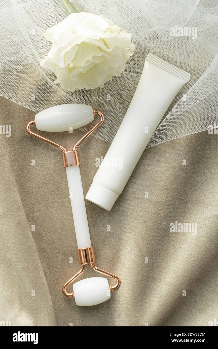 Une image apaisante mettant en vedette un tube cosmétique blanc et un rouleau facial sur tissu doux, complété par une fleur blanche délicate. Idéal pour la beauté et la peau Banque D'Images