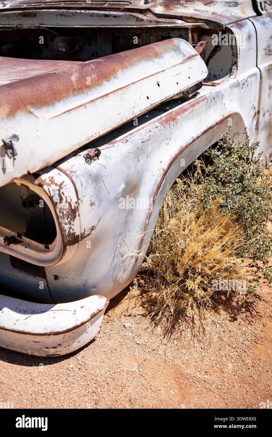 Gros plan d'une voiture rouillée abandonnée dans un paysage désertique, présentant du métal altéré, de la peinture décolorée et des broussailles envahissantes au milieu d'un terrain sablonneux sec. Bouchon Banque D'Images