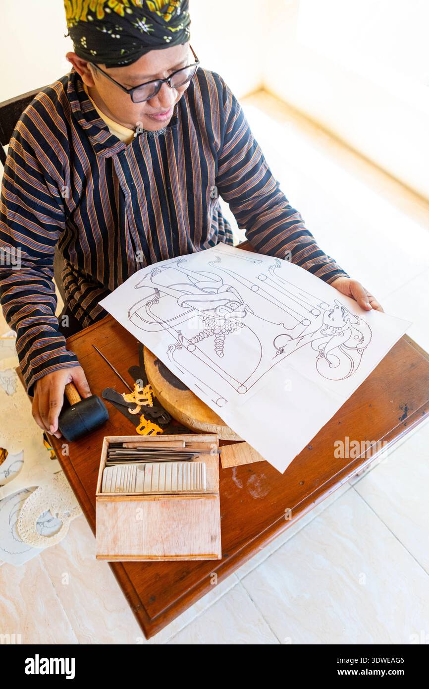 Artisan javanais souriant tout en affichant un dessin de marionnettes d'ombre pour Wayang Kulit, entouré d'outils de sculpture traditionnels sur une table en bois. Banque D'Images