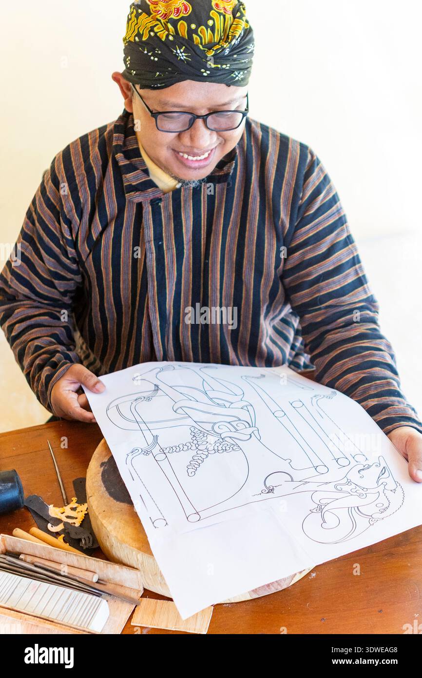 Artisan javanais souriant tout en affichant un dessin de marionnettes d'ombre pour Wayang Kulit, entouré d'outils de sculpture traditionnels sur une table en bois. Banque D'Images