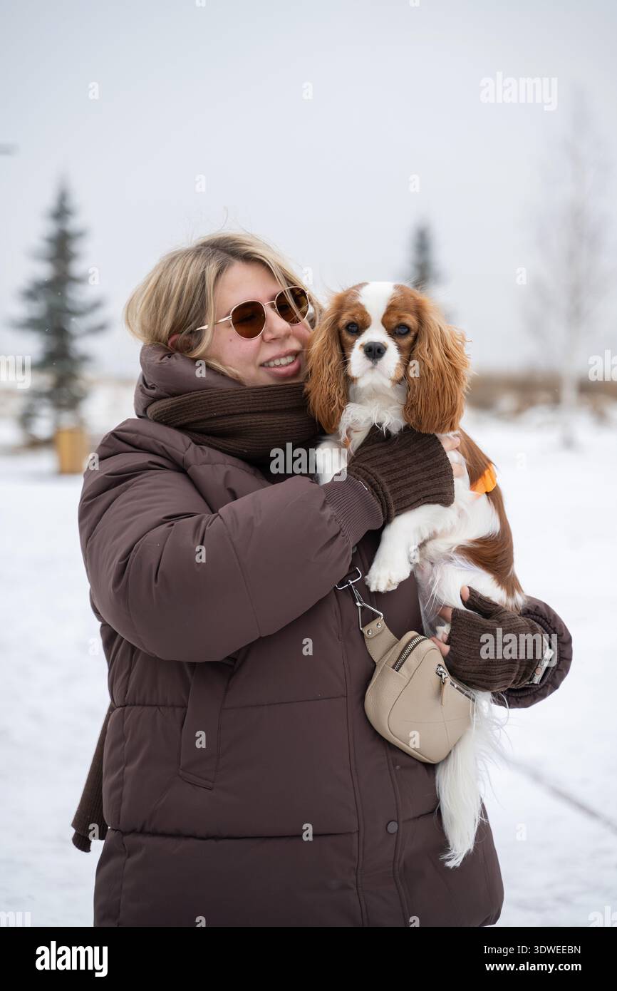 Une femme vêtue d'un manteau d'hiver tient un Cavalier King Charles Spaniel alors qu'elle profite d'une promenade dans la neige. La toile de fond pittoresque de l'hiver ajoute au m confortable et joyeux Banque D'Images