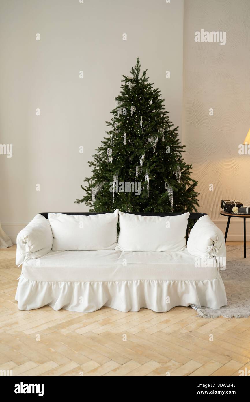 Salon minimaliste avec un canapé blanc et un sapin de Noël luxuriant orné de décorations élégantes. Un espace chaleureux et accueillant idéal pour les fêtes Banque D'Images