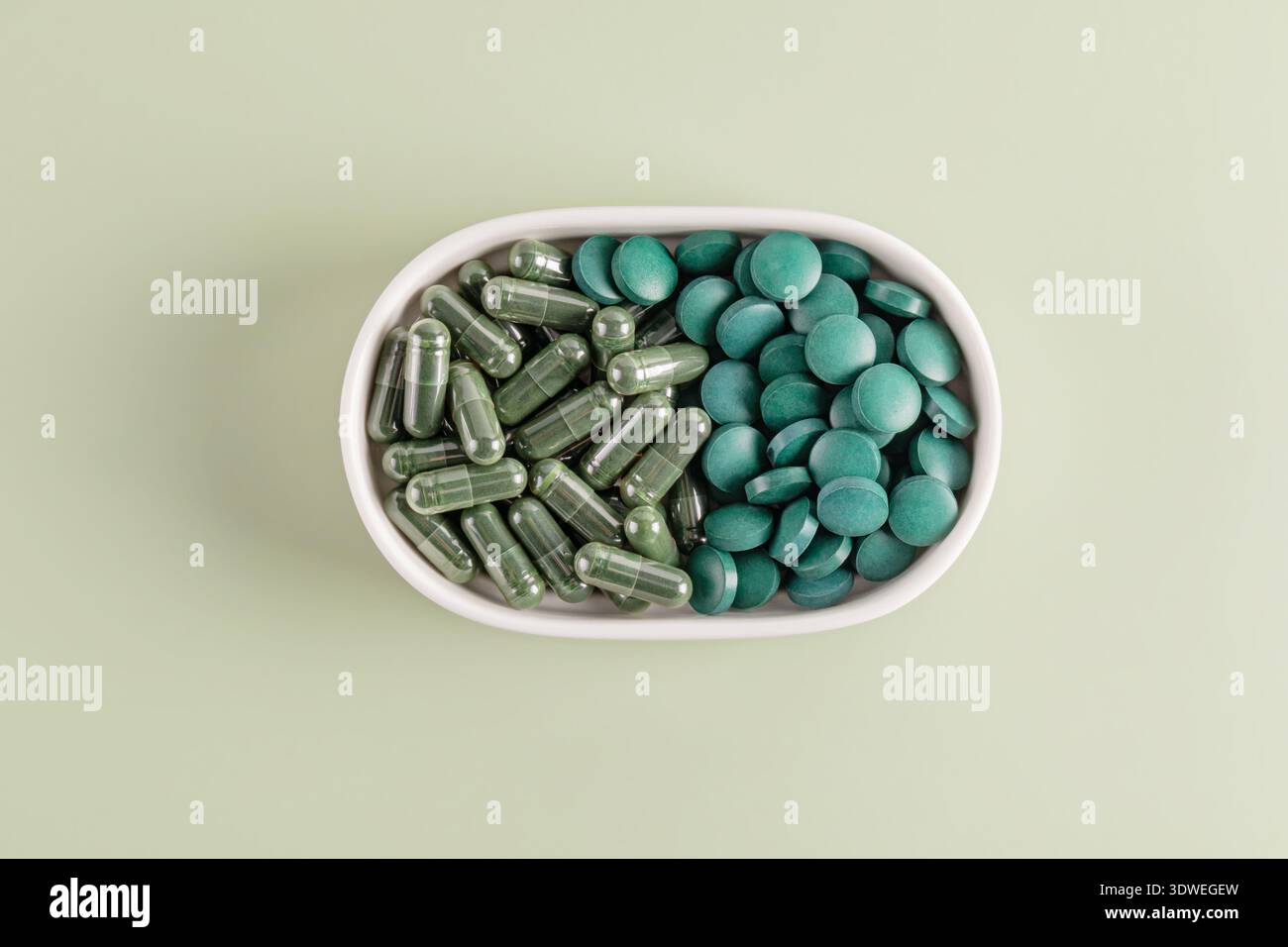 Vue de dessus des capsules et comprimés de spiruline affichés dans un plat ovale blanc sur un fond vert clair, soulignant leur couleur vibrante et leur texte lisse Banque D'Images