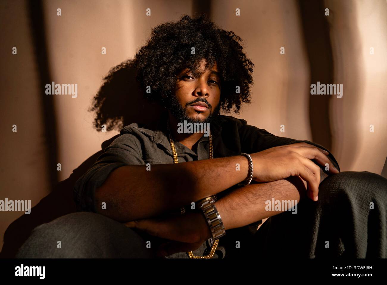 Jeune homme avec une coiffure afro assis en toute confiance dans un cadre de studio. Son expression est calme et introspective, avec des emphas d'éclairage monochromes Banque D'Images