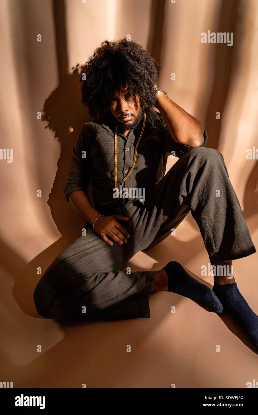 Un homme avec une coiffure afro portant une tenue élégante pose soigneusement dans un cadre de studio bien éclairé. Le jeu des ombres et de la lumière ajoute de la profondeur au Banque D'Images