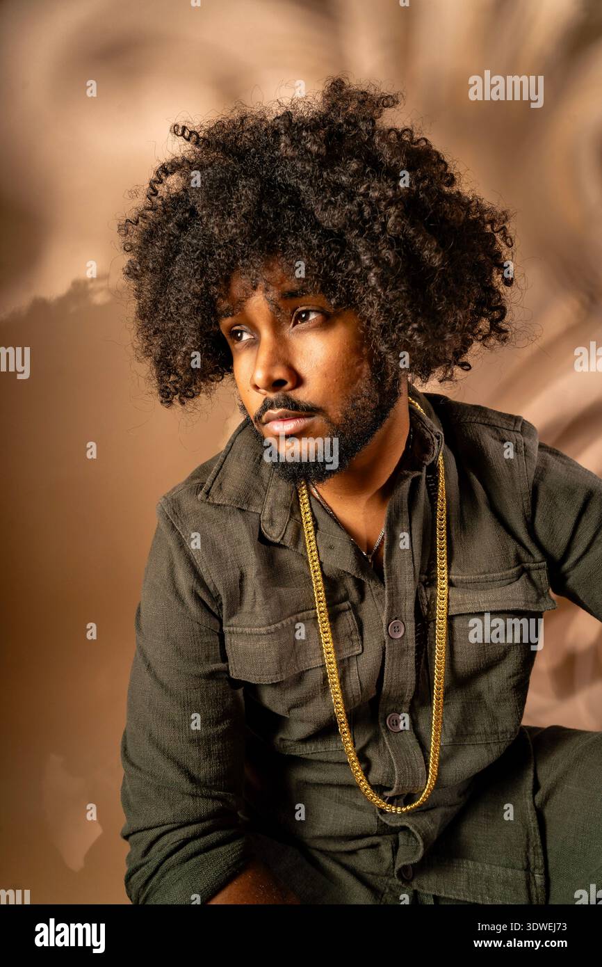 Portrait d'un homme avec une coiffure afro, orné d'une tenue à la mode, posant soigneusement dans un studio. Le fond est légèrement flou, enhancin Banque D'Images