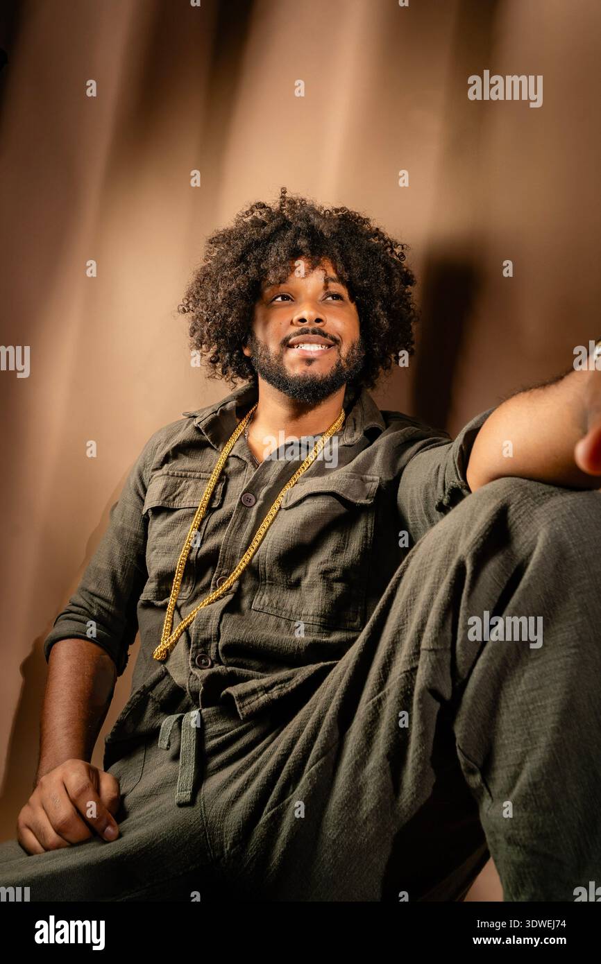 Un homme détendu avec une coiffure afro est assis dans un cadre de studio, portant une tenue décontractée et une chaîne en or. Le fond brun chaud complète son con Banque D'Images