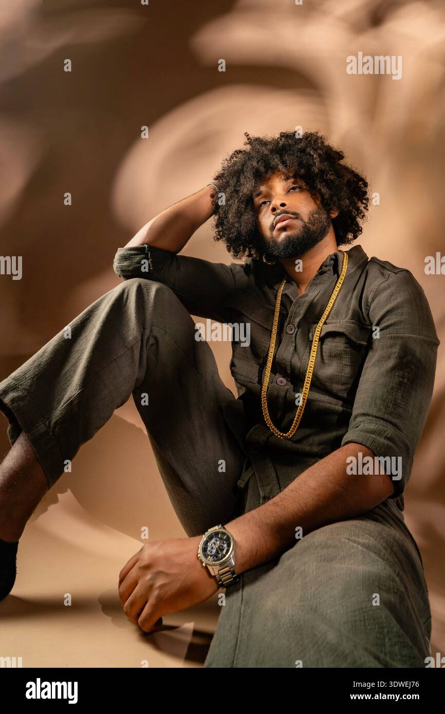 Un homme avec une coiffure afro s'assoit soigneusement dans un cadre de studio. Il porte une tenue décontractée, accessoirisée d'une chaîne et d'une montre en or, exsudant un rela Banque D'Images