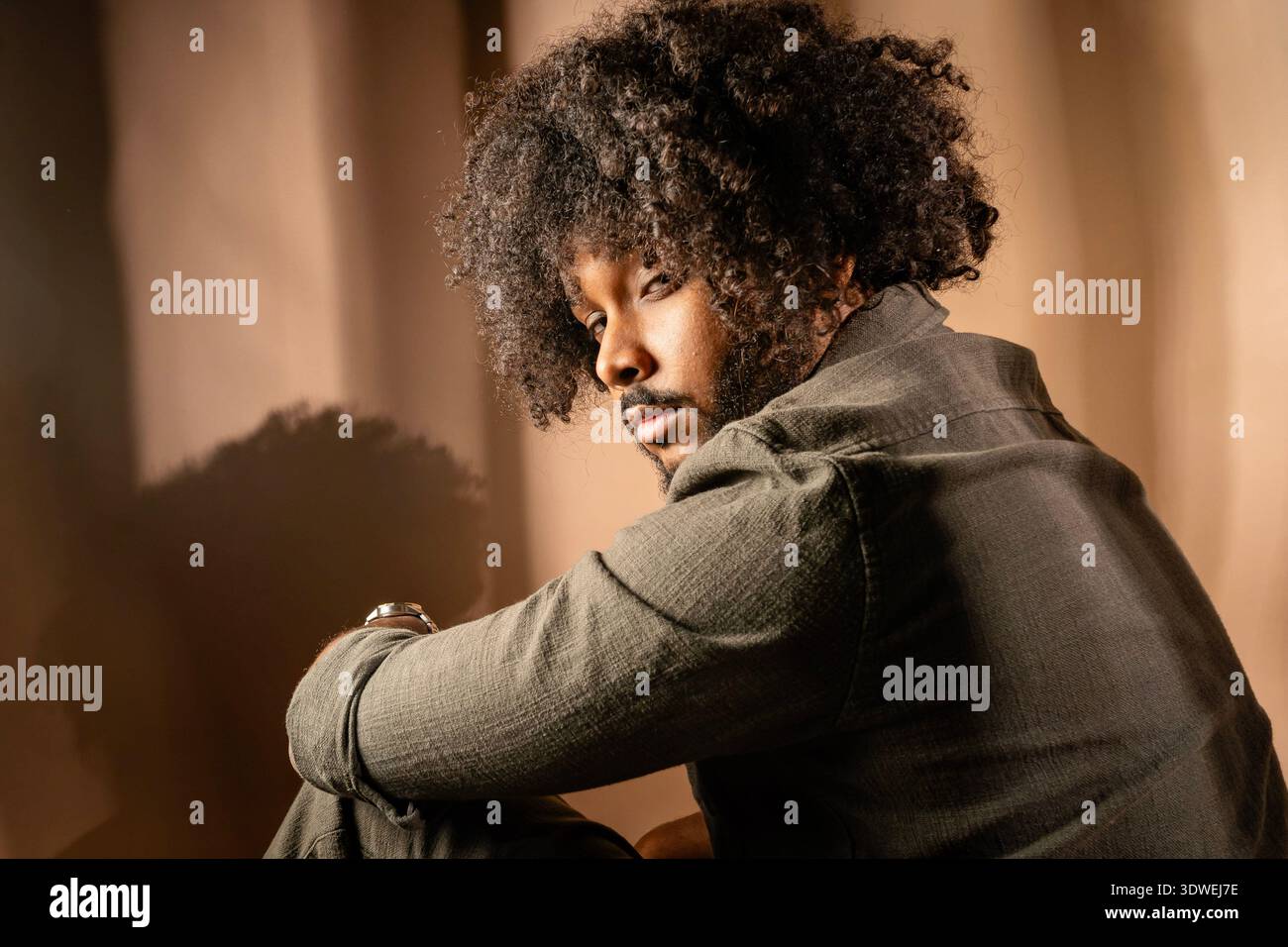 Un homme avec une coiffure afro pose avec confiance dans un cadre de studio. L'éclairage chaleureux met en valeur sa tenue décontractée et son expression réfléchie, créant Banque D'Images