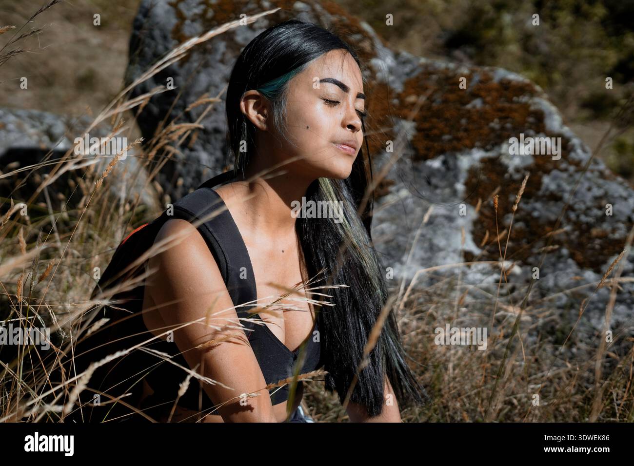 Jeune femme routard prend une pause de la randonnée, assis parmi les hautes herbes et profiter de la lumière chaude du soleil sur son visage, avec un terrain rocheux dans le b. Banque D'Images