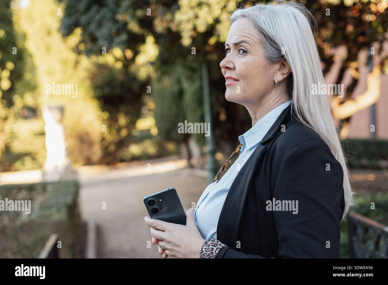 Femme d'affaires senior portant une veste noire et une chemise bleue tient un smartphone et regarde loin tout en pensant à quelque chose dans un parc pendant un Banque D'Images