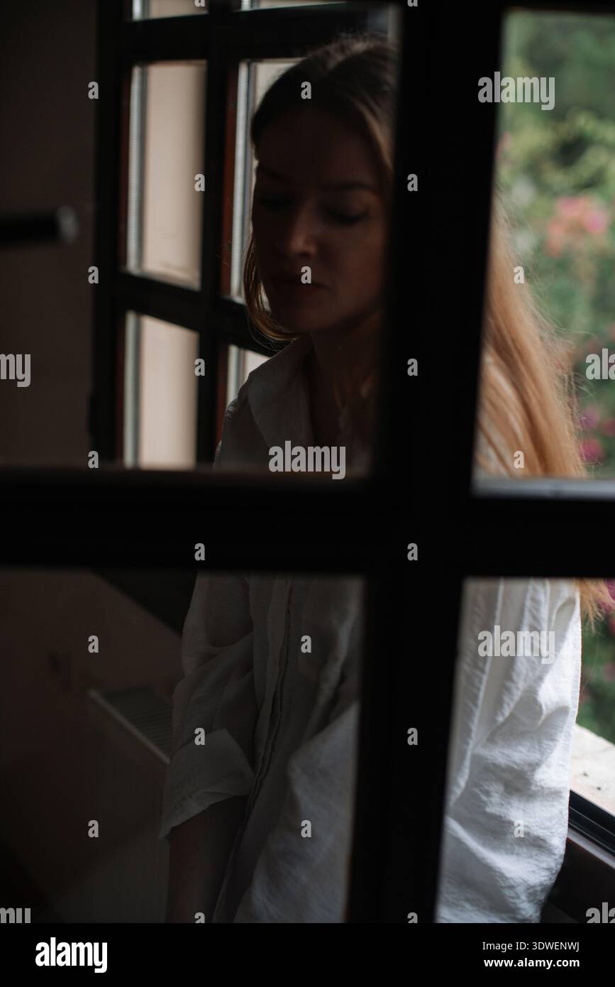 Une femme réfléchie se tient près d'une fenêtre ouverte, son visage à moitié éclairé par la lumière douce et naturelle. Elle est capturée dans un moment contemplatif, Surround Banque D'Images