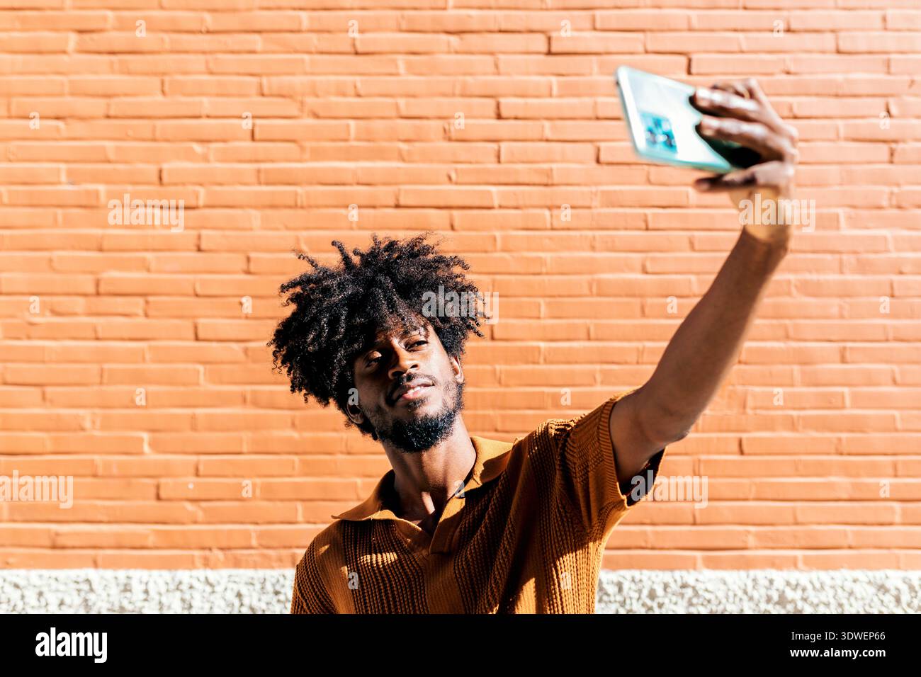Un homme avec une coiffure afro prend un selfie contre un mur de briques orange ensoleillé. Il porte une chemise texturée marron et semble détendu et confiant Banque D'Images
