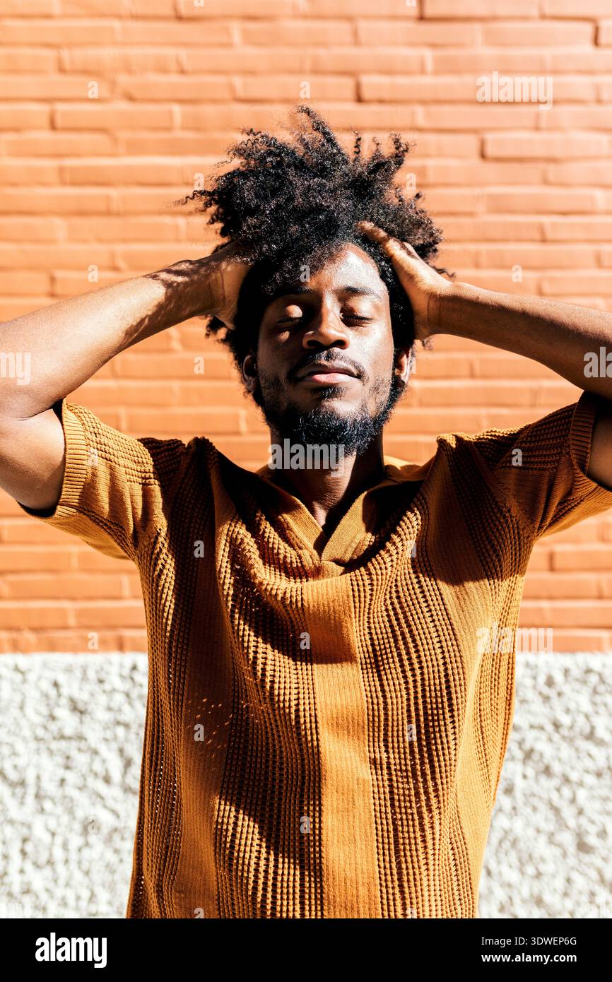 Un homme noir avec une coiffure afro se tient confortablement à l'extérieur, se prélassant à la lumière du soleil contre un mur de briques texturées. Ses yeux sont fermés, véhiculant un se Banque D'Images
