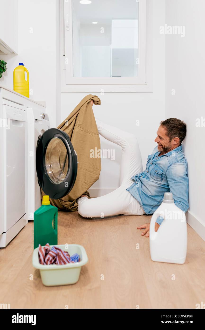 Homme souriant assis sur le sol d'une buanderie moderne, manipulant ludique des vêtements devant une machine à laver, entouré de bouteilles de détergent A. Banque D'Images