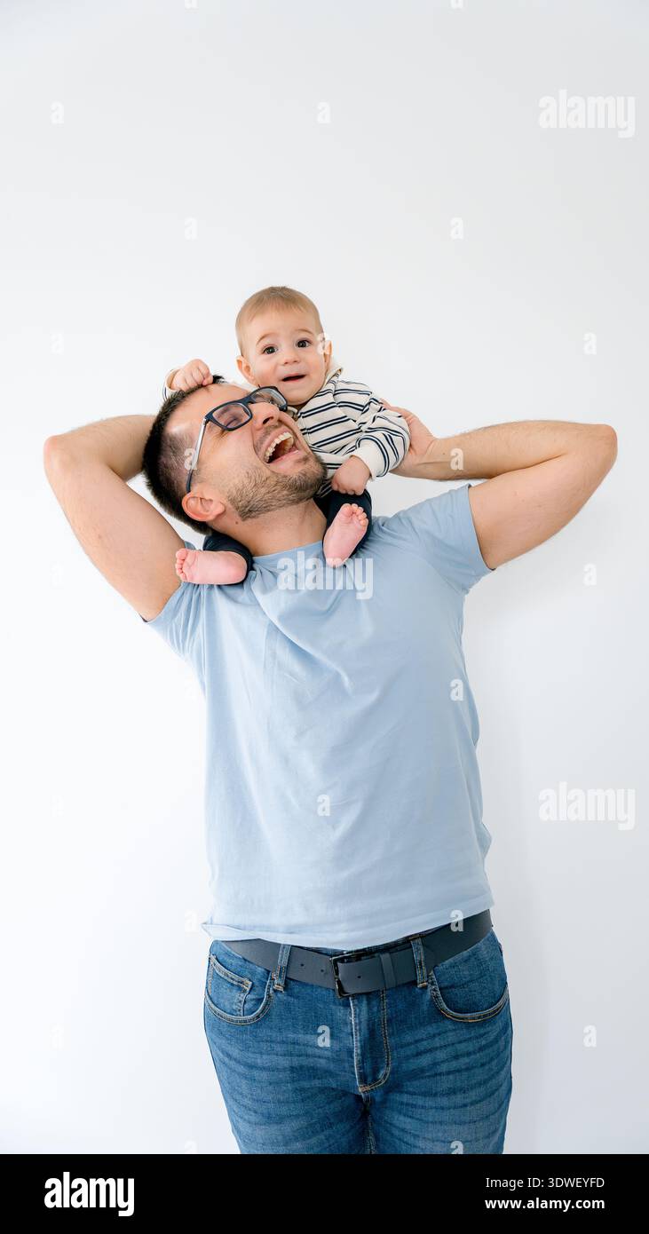 Un père joyeux avec des lunettes tient son bébé en l'air, souriant vigoureusement alors qu'ils partagent un moment ludique. Le bébé, vêtu de rayures, profite de la balade sur h. Banque D'Images