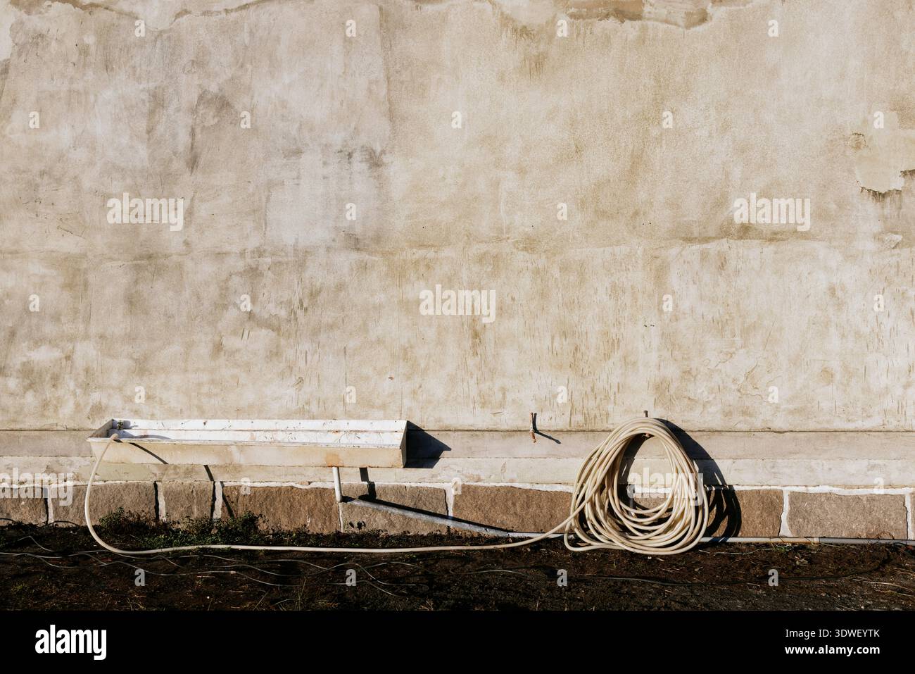 Un mur extérieur vieilli et texturé avec un tuyau d'arrosage enroulé sur le sol, projetant des ombres douces. Les tons discrets et la lumière naturelle créent un atmos rustique Banque D'Images