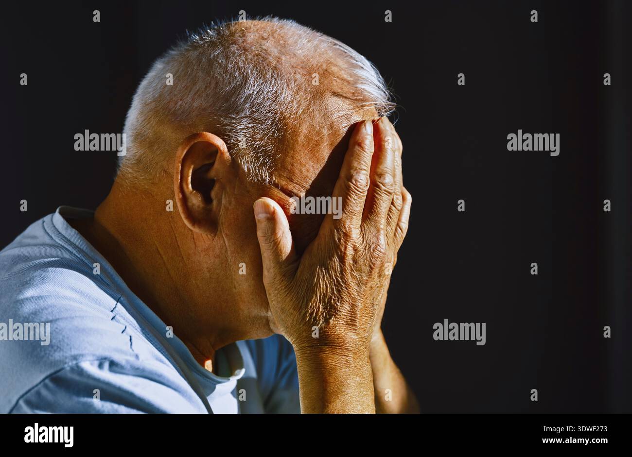 Un homme âgé aux cheveux gris est assis de profil avec ses mains obscurcissant son visage, capturant les détails et la texture de sa peau ridée, reflétant un Banque D'Images