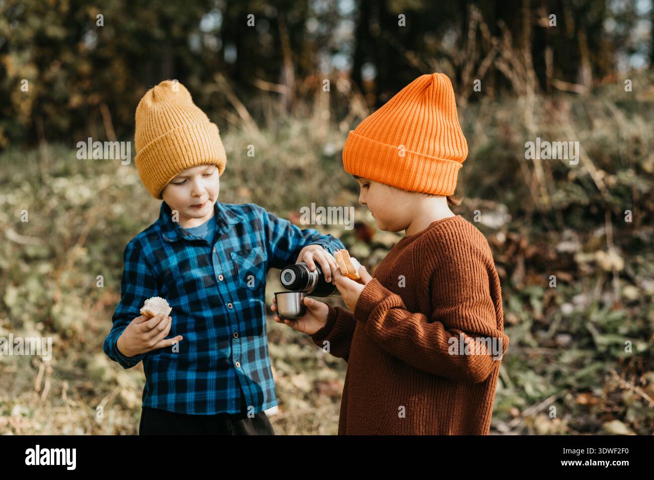 Deux enfants explorent une forêt en automne, s'engageant dans un mini voyage. Portant des chapeaux confortables et des pulls, ils partagent un verre, immergés dans la beauté et WO Banque D'Images