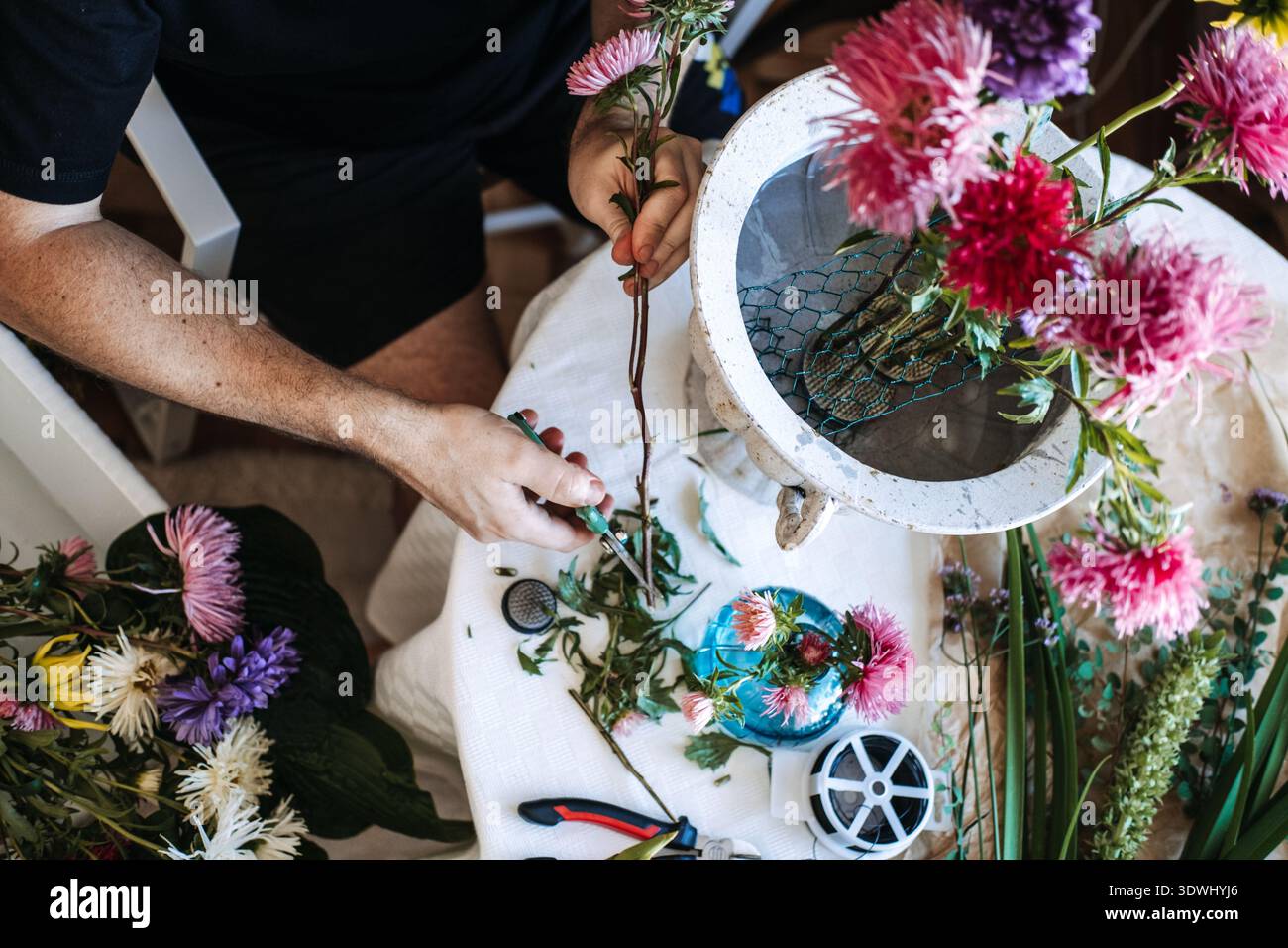 Les mains utilisent des ciseaux floraux pour couper la tige de fleur sur une table blanche avec diverses fleurs et urne. verdure détox numérique, plantes abaissant le cortisol, nature Banque D'Images