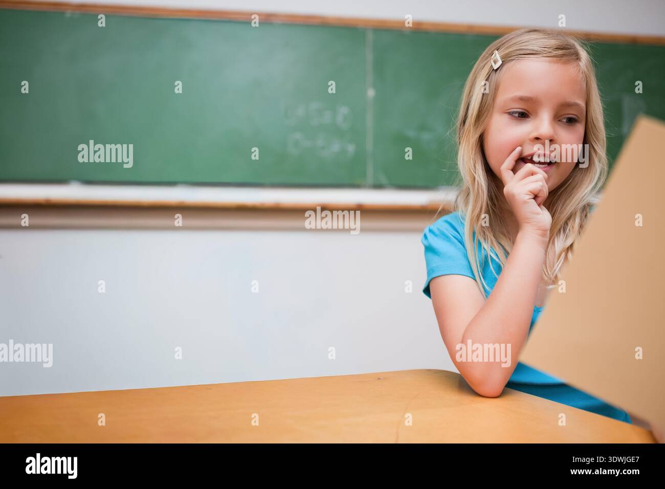 Fille assise au bureau de la classe tenant du papier bronzé portant un panneau supérieur bleu derrière, espace de copie. Tableau noir, étudiant, concentration, éducation, étude, blonde Banque D'Images
