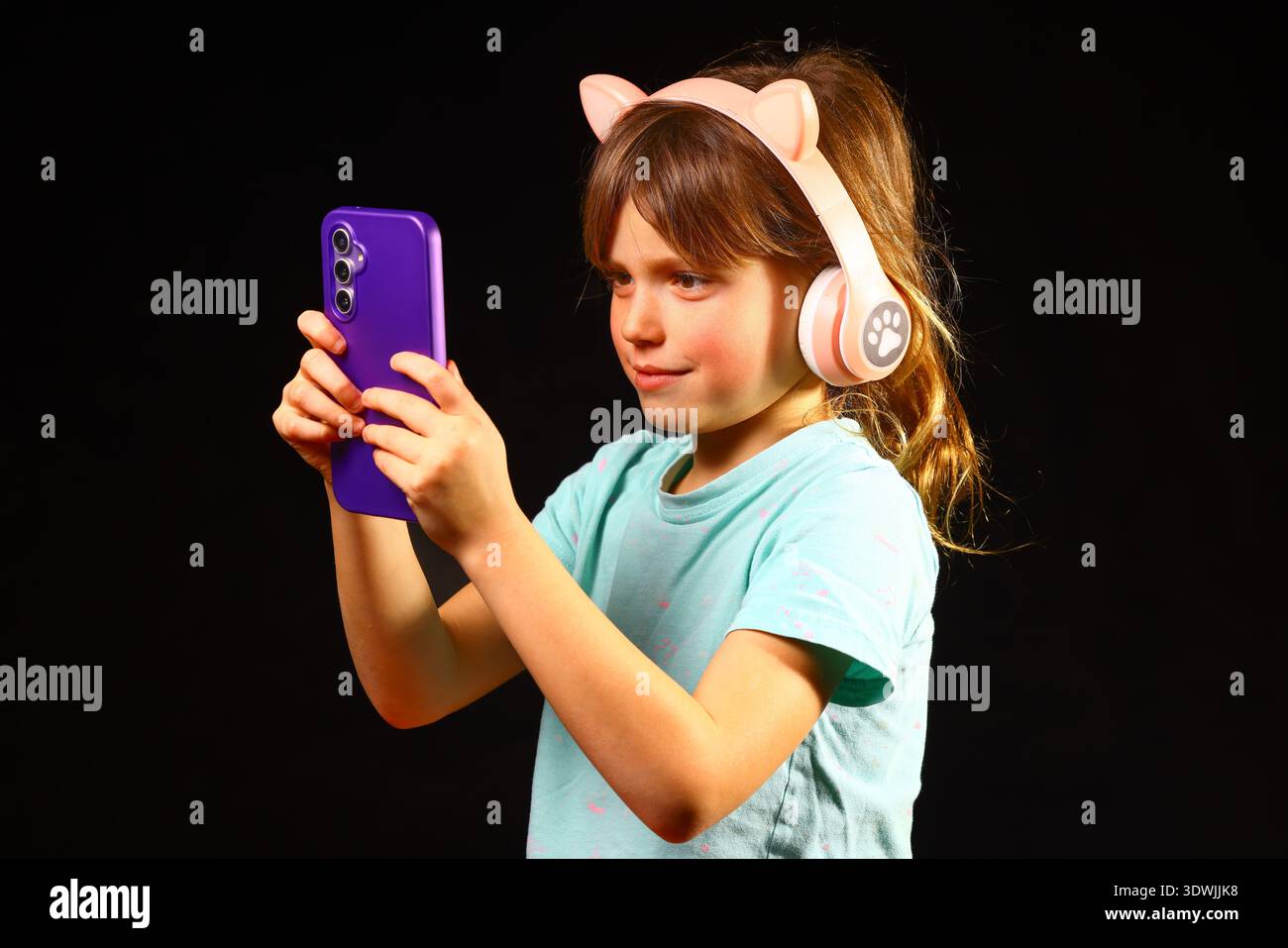 Enfant utilisant un smartphone tout en portant un casque d'oreille de chat rose dans un portrait de studio sombre, représentant l'utilisation de la technologie par les enfants, l'apprentissage numérique, le mobil moderne Banque D'Images