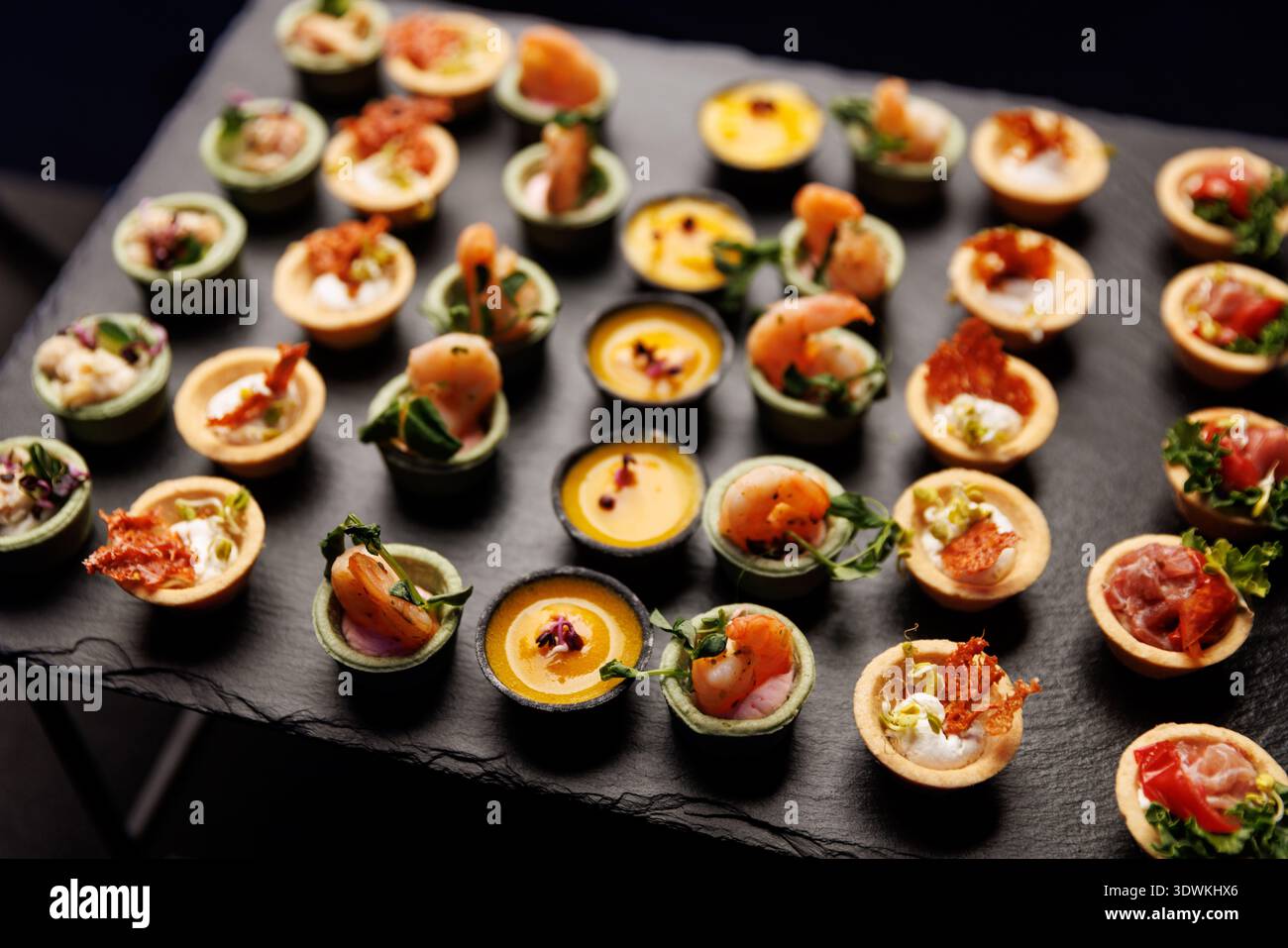 Un éventail de petites coquilles de pâtisserie remplies de crevettes, purées et herbes sont soigneusement disposées sur un plateau de pierre noire. Banque D'Images