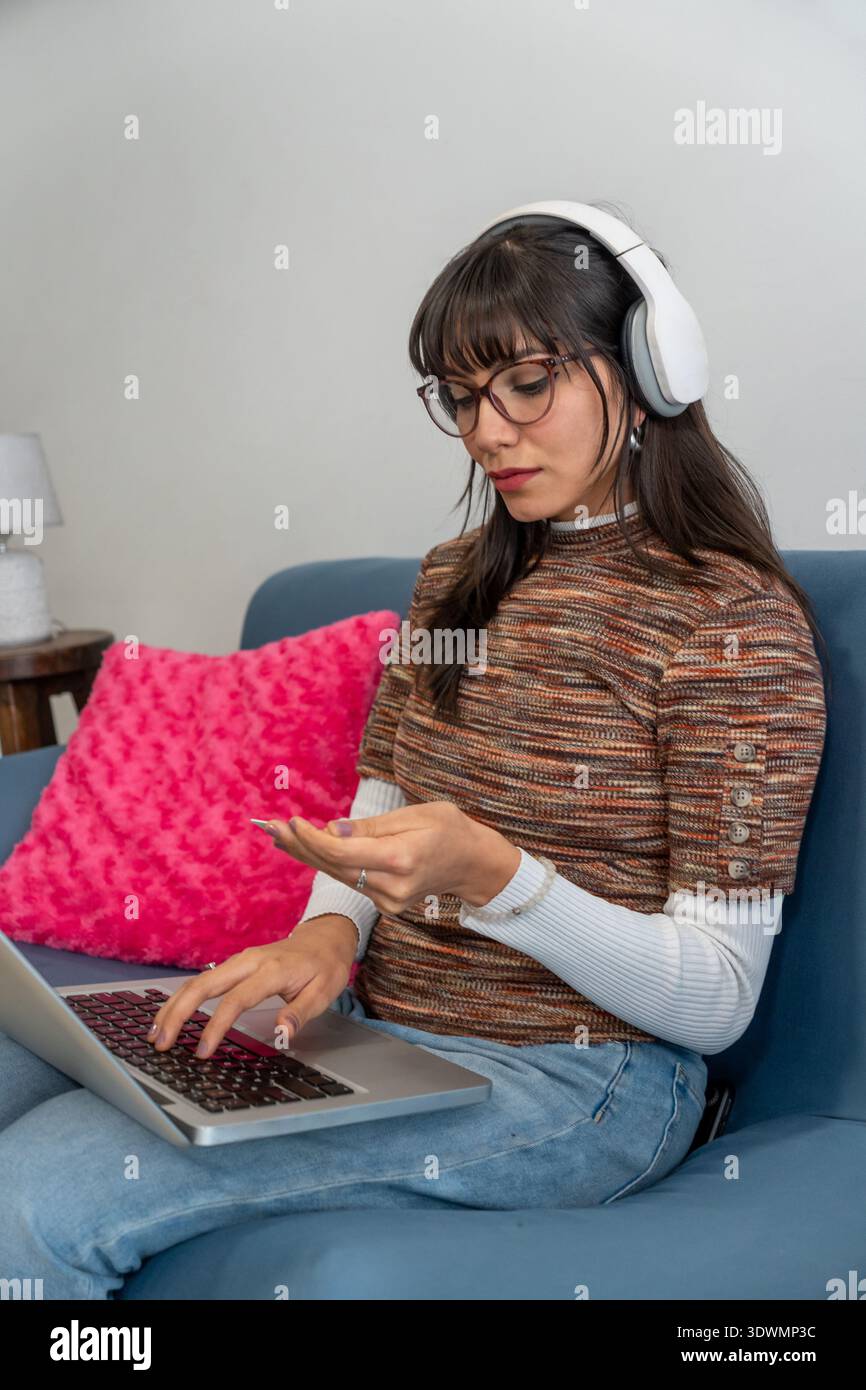 Jeune femme portant des écouteurs et des lunettes travaillant à distance depuis un canapé, tapant sur un ordinateur portable Banque D'Images