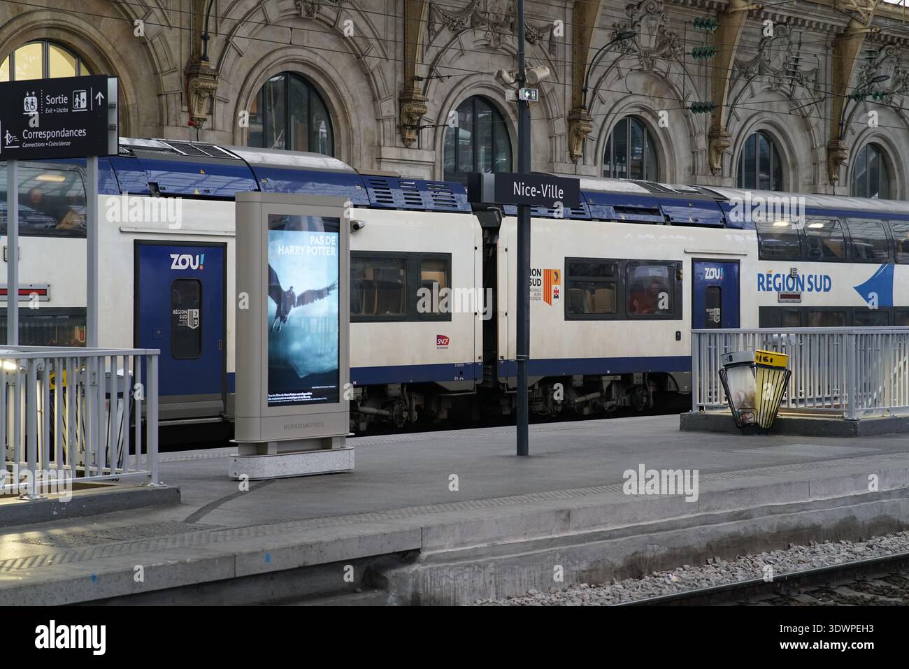 Le train régional bleu et blanc du Zou repose sur la plate-forme historique de la Gare de Nice ville sous de grandes arches en pierre et un panneau d'affichage numérique lumineux. Banque D'Images