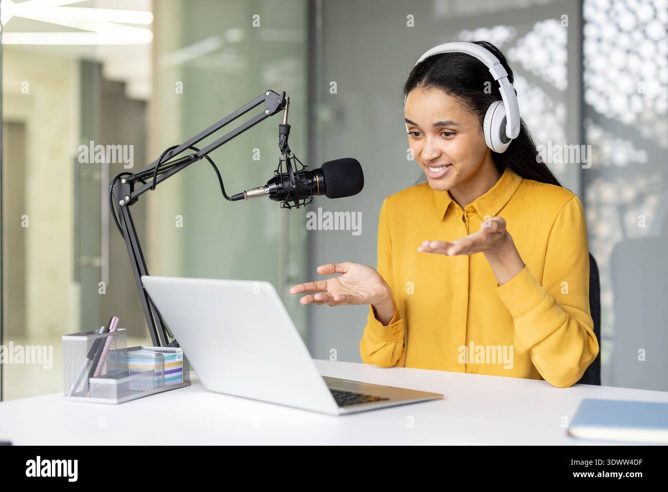 Jeune femme biraciale enregistrant un podcast, portant des écouteurs et parlant dans un microphone professionnel tout en regardant un ordinateur portable, partageant du contenu dans un espace de travail moderne Banque D'Images