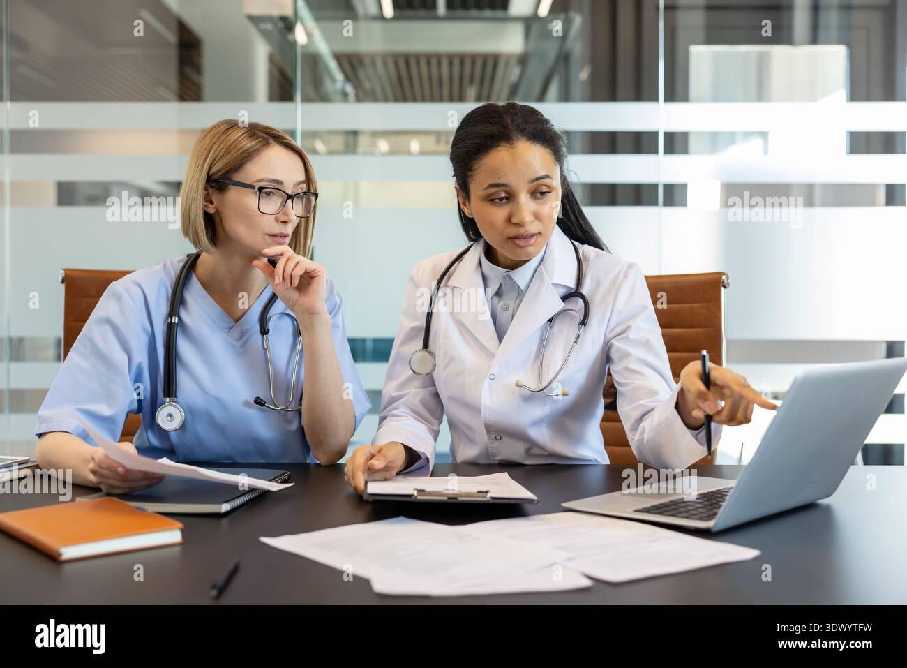 Deux professionnels de la santé féminins divers sont activement engagés dans une discussion, examinant des données importantes sur un ordinateur portable et des documents tout en travaillant ensemble dans une salle de réunion moderne de bureau d'hôpital Banque D'Images