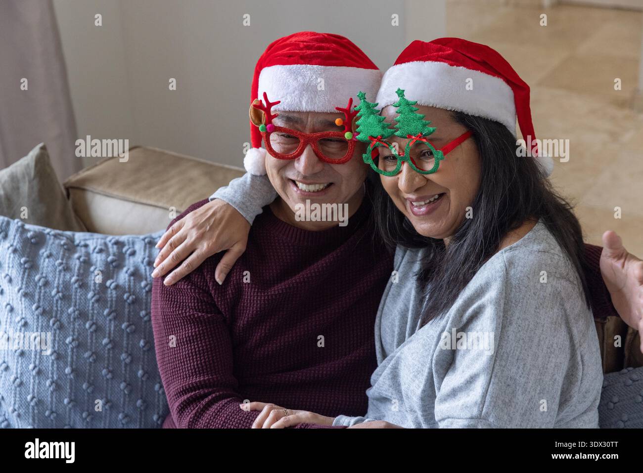 Couple senior diversifié souriant tout en portant des chapeaux de Père Noël et des lunettes de fête sur le canapé dans le salon. Festif, célébration, compagnie, chaleur, vacances, Joy, joyeuse || autorisation du modèle Banque D'Images