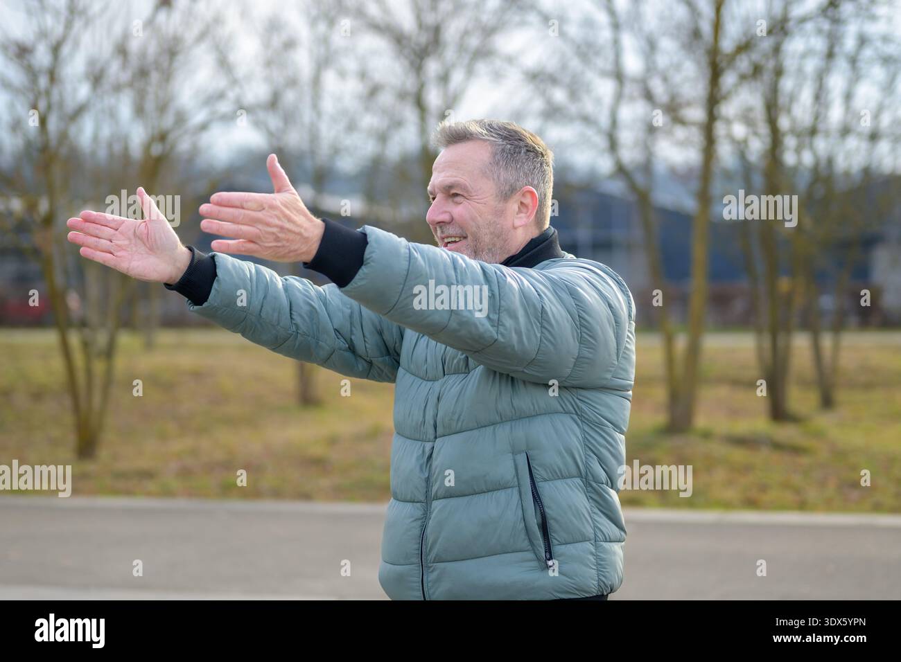 Homme mature souriant dans une veste verte étirant ses bras à l'extérieur dans un parc, profitant de l'air frais et de l'exercice léger par une journée ensoleillée. Banque D'Images