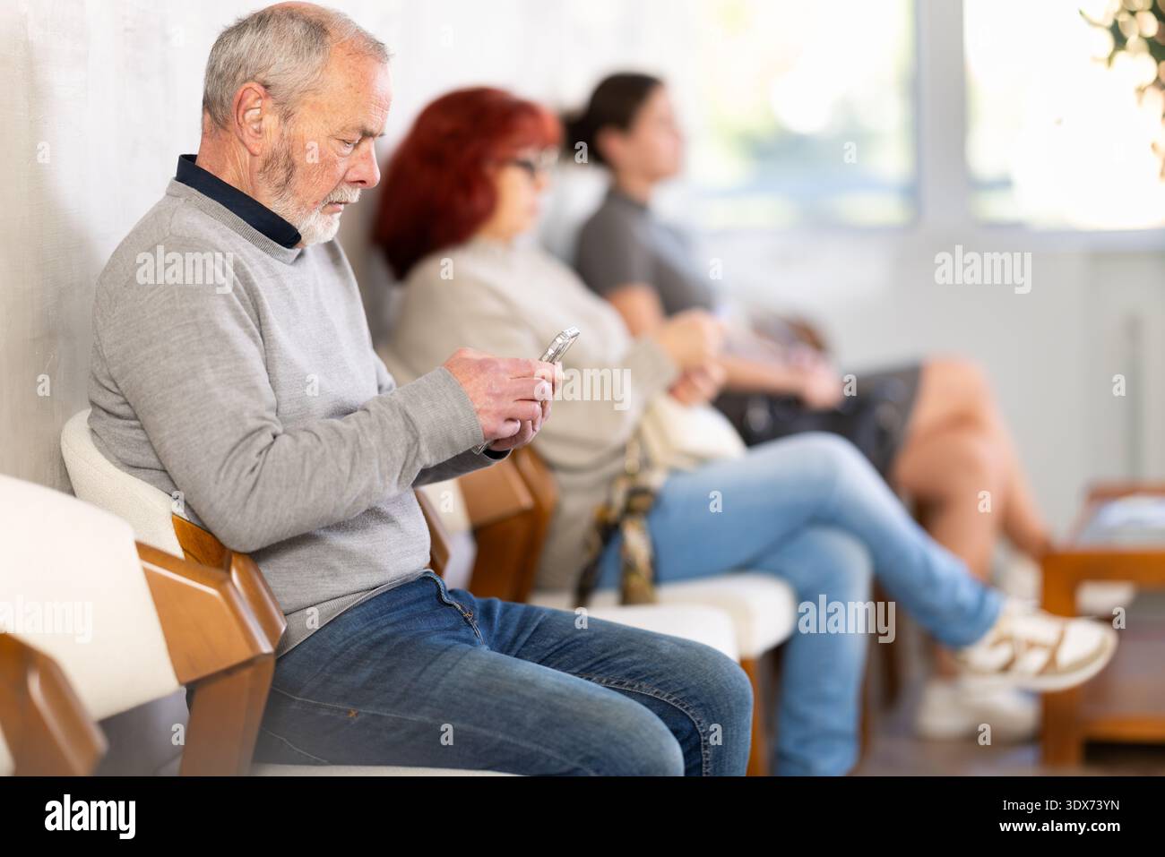 Homme âgé assis dans la salle d'attente clinique défilant téléphone mobile Banque D'Images