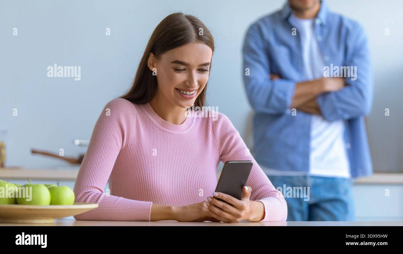 Femme sourit tout en utilisant smartphone dans la cuisine avec l'homme debout derrière elle Banque D'Images