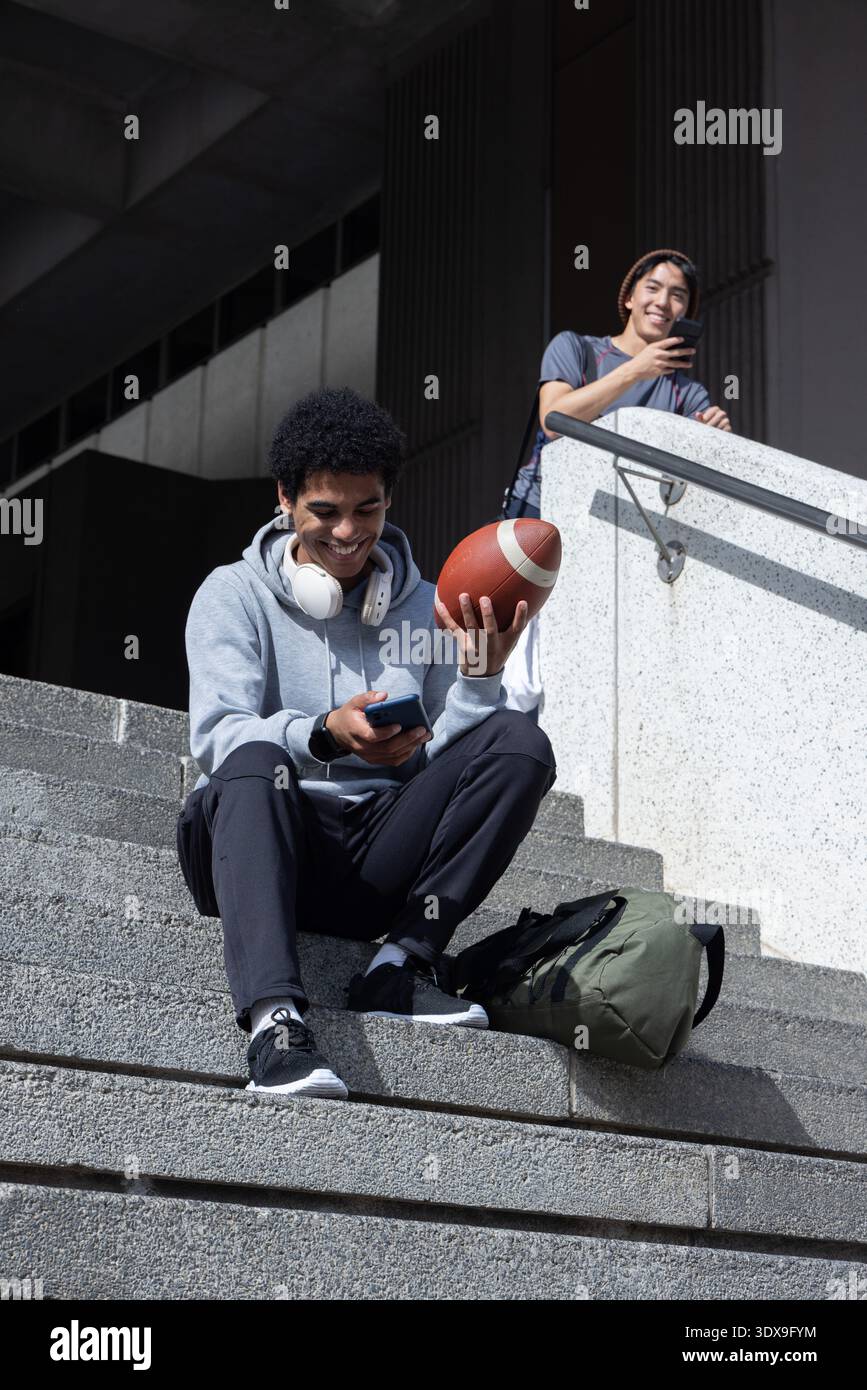 Afro-américains et asiatiques adolescents hommes assis sur le campus étapes en béton tenant le football, téléphones Banque D'Images