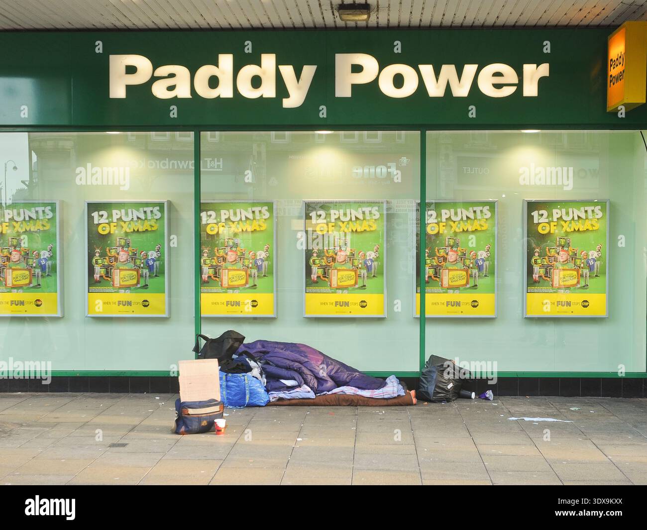 Un sans-abri dort dans une porte devant un magasin de Paris Paddy Power, placé sur un trottoir public. La scène présente le contraste entre la literie de fortune de l'individu et la marque vert vif de l'établissement de jeu. Ces boutiques de Paris sont fréquemment situées dans des zones de rue avec une fréquentation importante, où les entrées en retrait fournissent un abri temporaire contre les intempéries. Banque D'Images