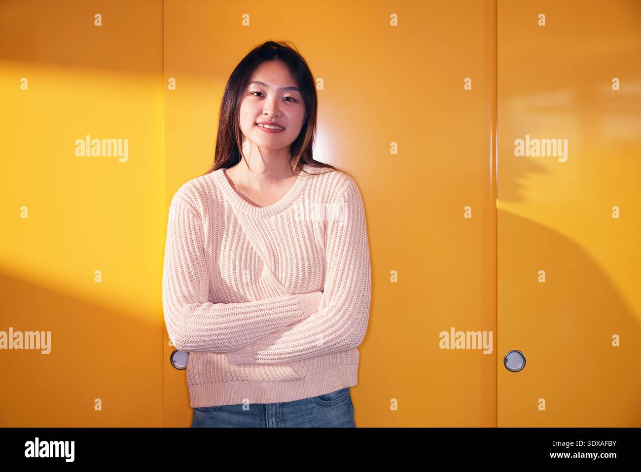 Portrait d'une jeune femme souriante debout avec les bras croisés contre le mur jaune Banque D'Images