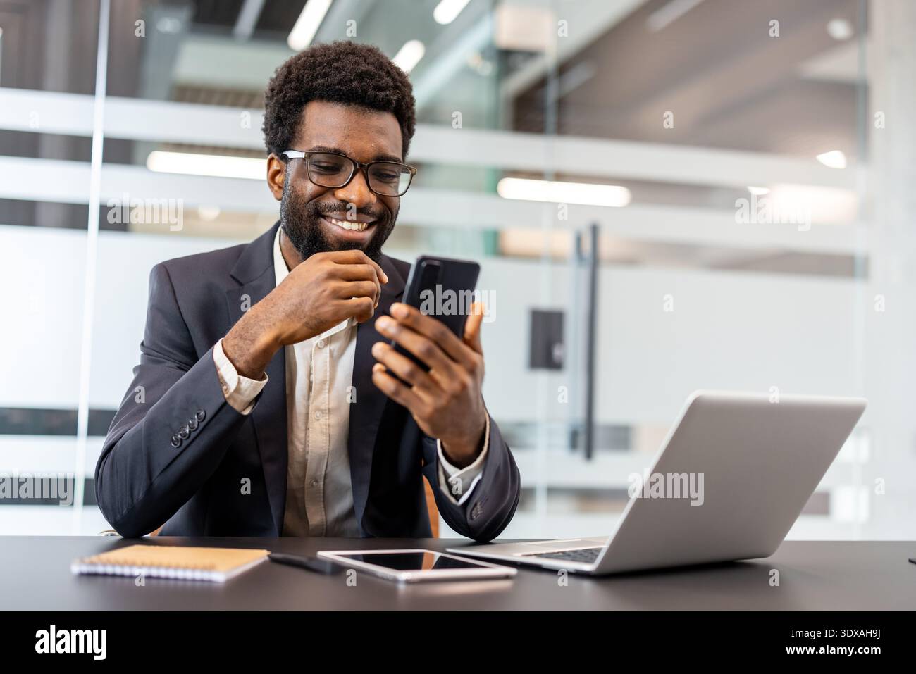 Homme d'affaires souriant naviguant smartphone à un bureau avec ordinateur portable ouvert dans un bureau moderne, à l'air positif et connecté tout en travaillant à distance et en restant productif et professionnel Banque D'Images