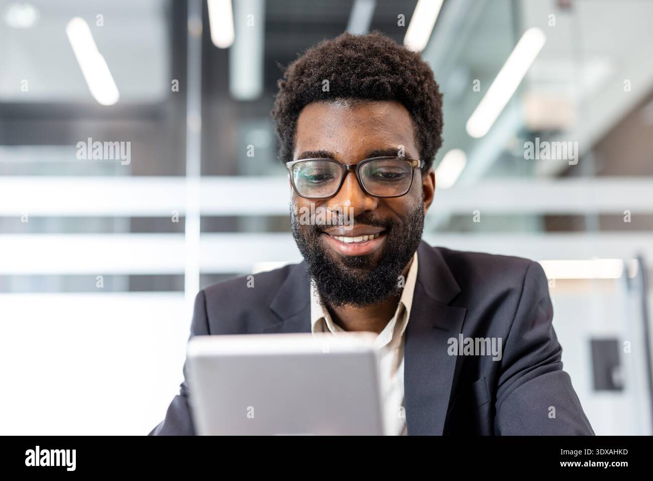 Jeune homme portant des lunettes et un costume de travail en toute confiance travaillant et souriant tout en utilisant une tablette numérique pour la communication professionnelle dans un espace de travail contemporain lumineux Banque D'Images