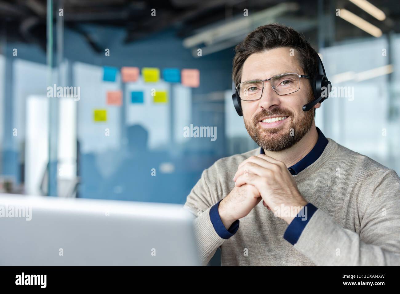 Homme barbu heureux travaillant dans un bureau moderne, portant un casque avec un microphone et des lunettes, fournissant un support client en ligne ou participant à une visioconférence Banque D'Images