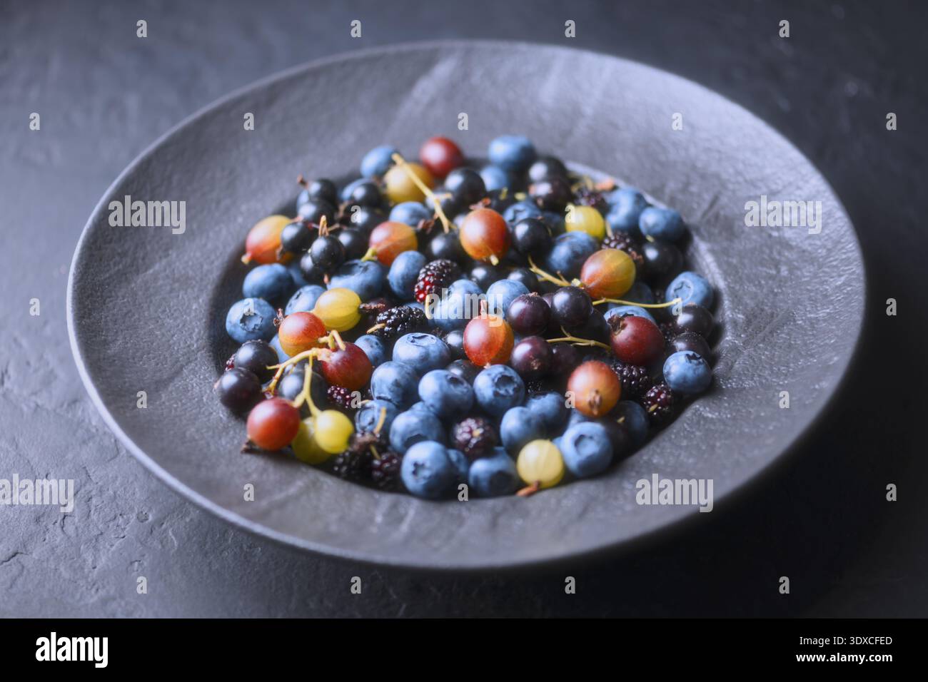 Baies d'été mélangées dans un bol gris, composition de vue de dessus sur une table sombre. Myrtilles, mûres et groseilles à maquereau, récolte biologique Banque D'Images