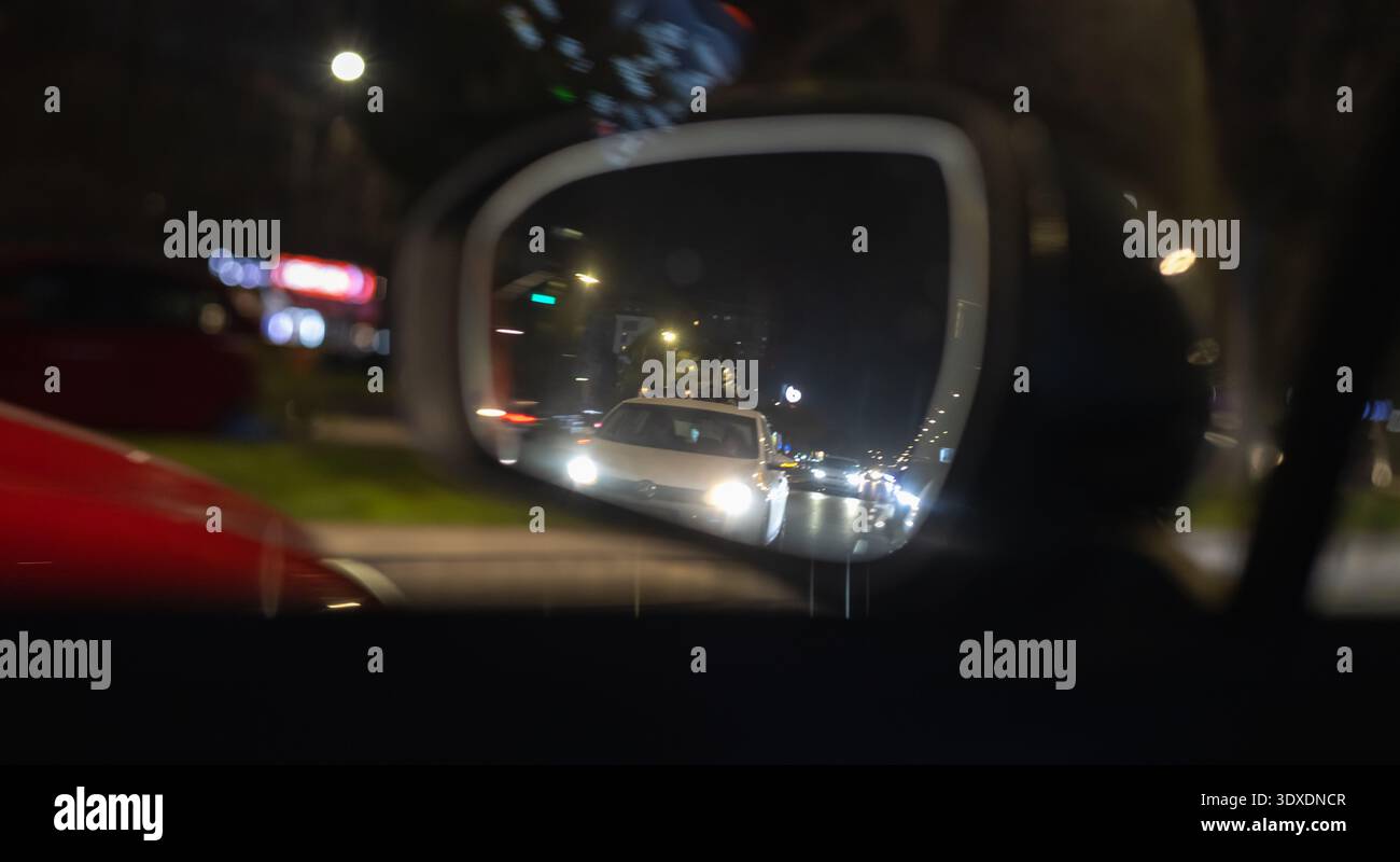 nuit cinématographique filmée à travers un rétroviseur latéral de voiture montrant un véhicule blanc avec des phares brillants dans la circulation, bokeh urbain et lumières de ville floues dans le b Banque D'Images
