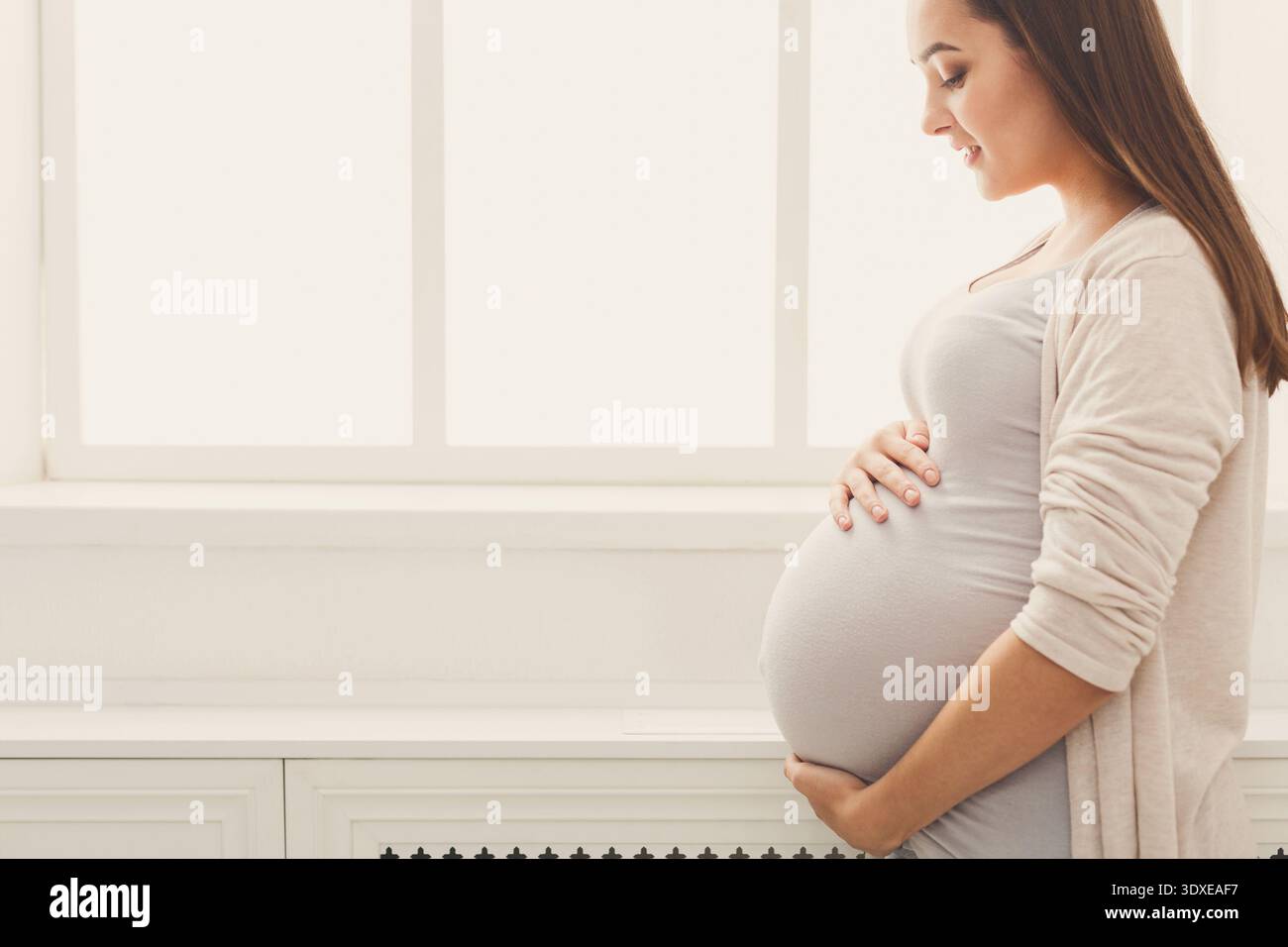 Femme enceinte pose près d'une fenêtre avec ses mains reposant sur son ventre dans un cadre calme Banque D'Images