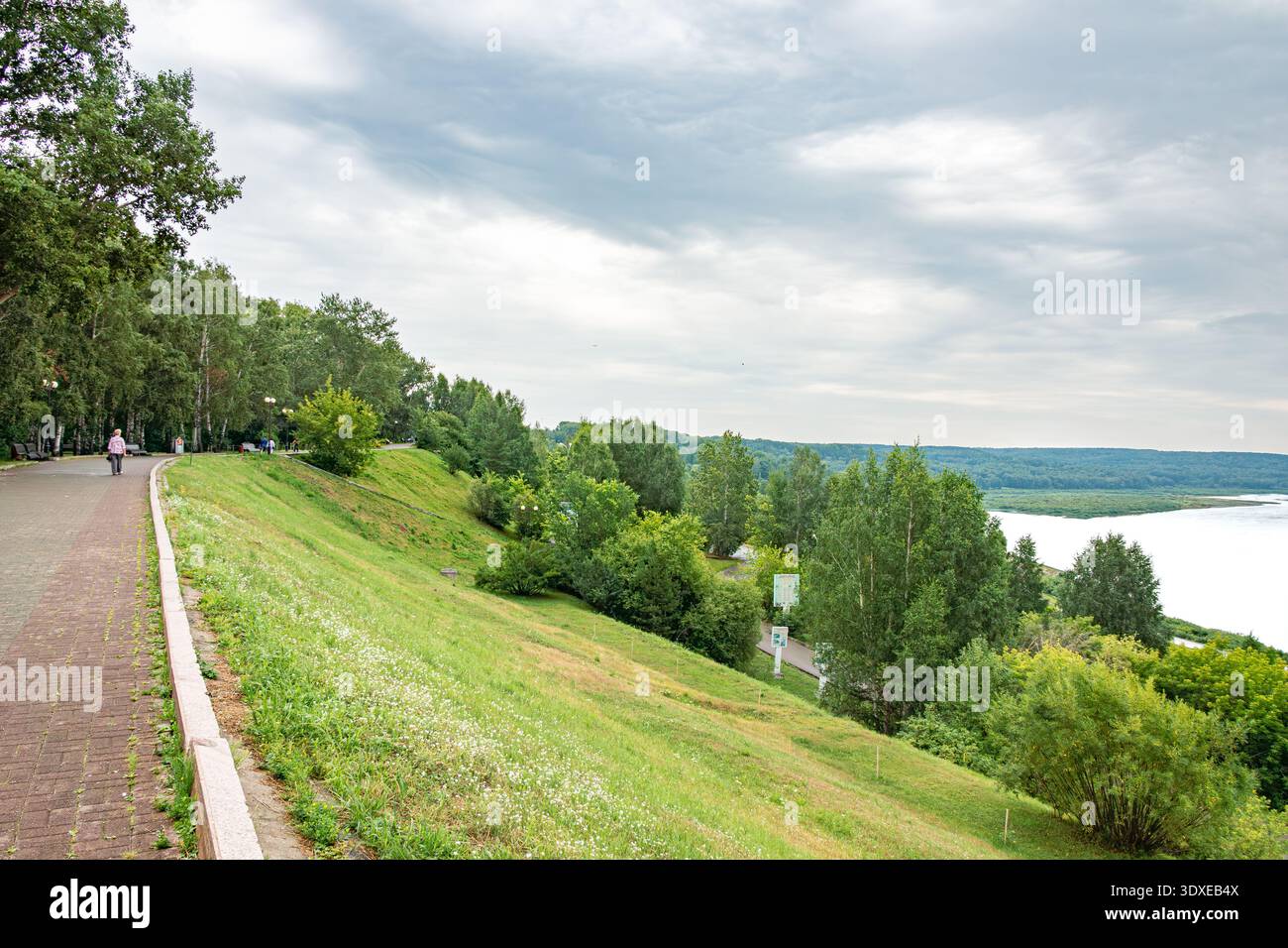 Tomsk, Russie - 19 juillet 2025. Une passerelle pavée longe la rive, bordée de bordures et de forêts mixtes de feuillus. Pente douce couverte de veget de prairie Banque D'Images