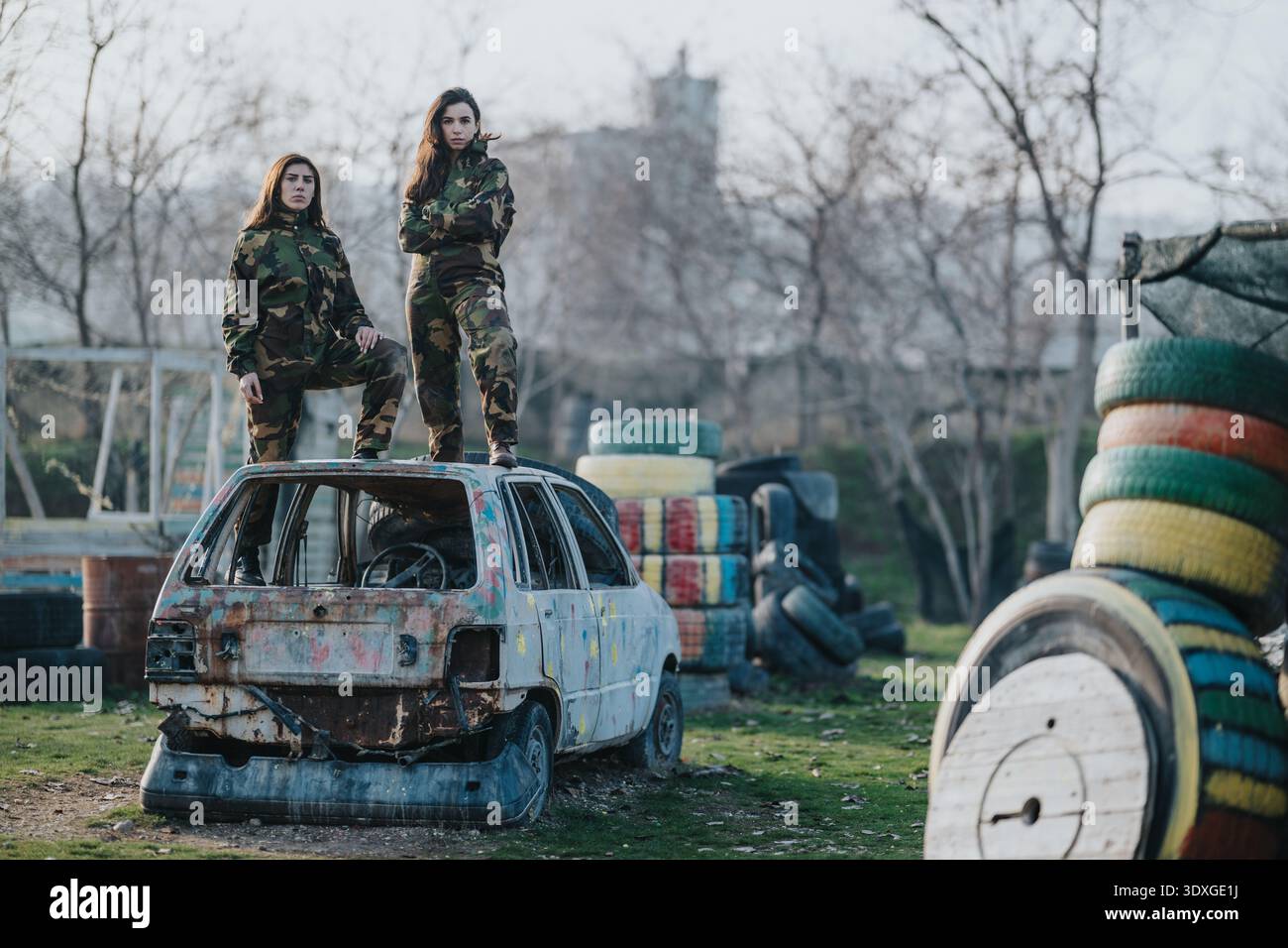 Deux femmes en camouflage se tiennent debout sur une voiture rouillée et abandonnée dans un environnement de casse-croûte en plein air encombré Banque D'Images