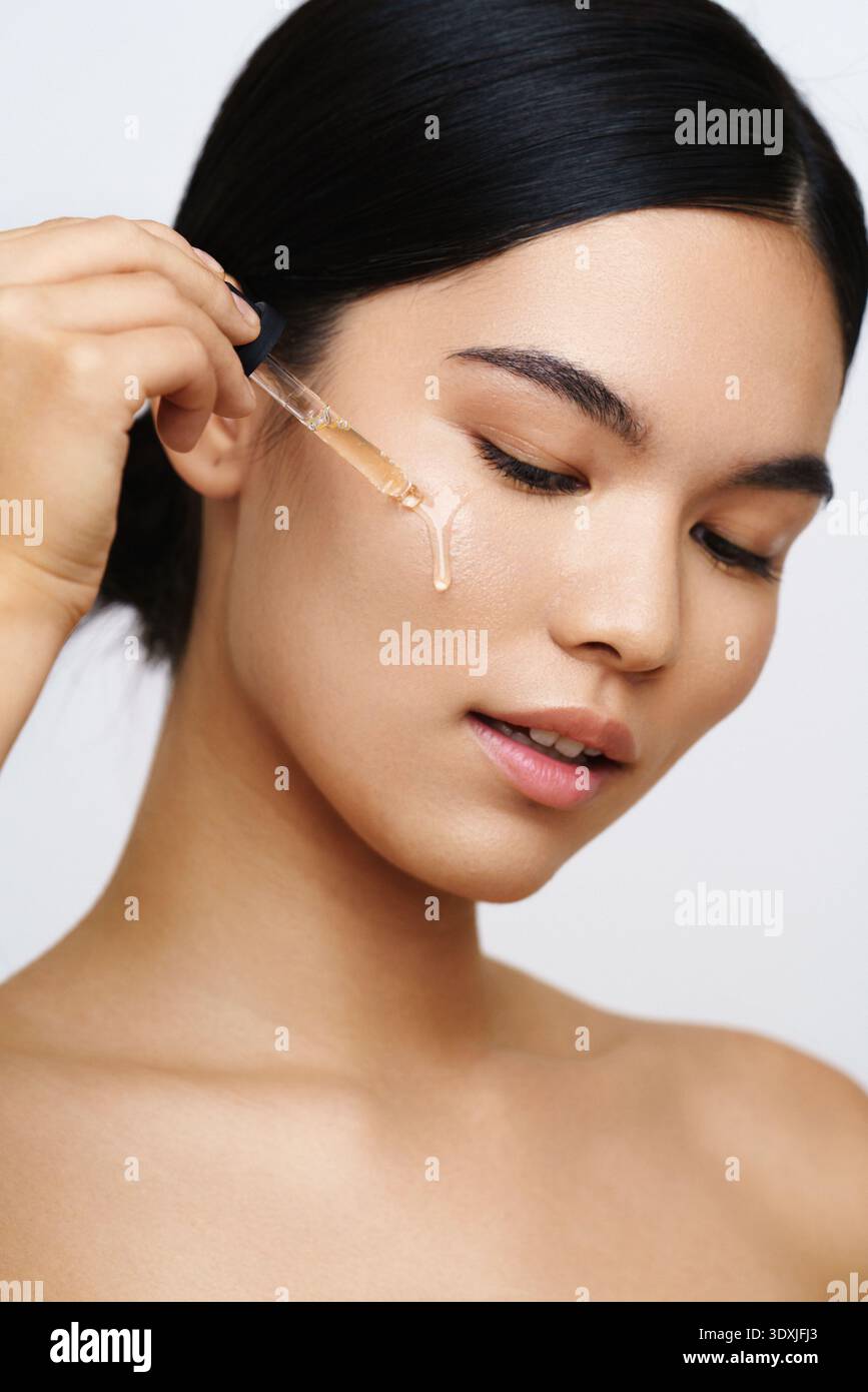 Une femme applique une goutte de sérum sur sa joue avec un compte-gouttes, mettant en valeur une routine de soins de la peau. L'image met en évidence les concepts de beauté et d'auto-soin dans un cl Banque D'Images