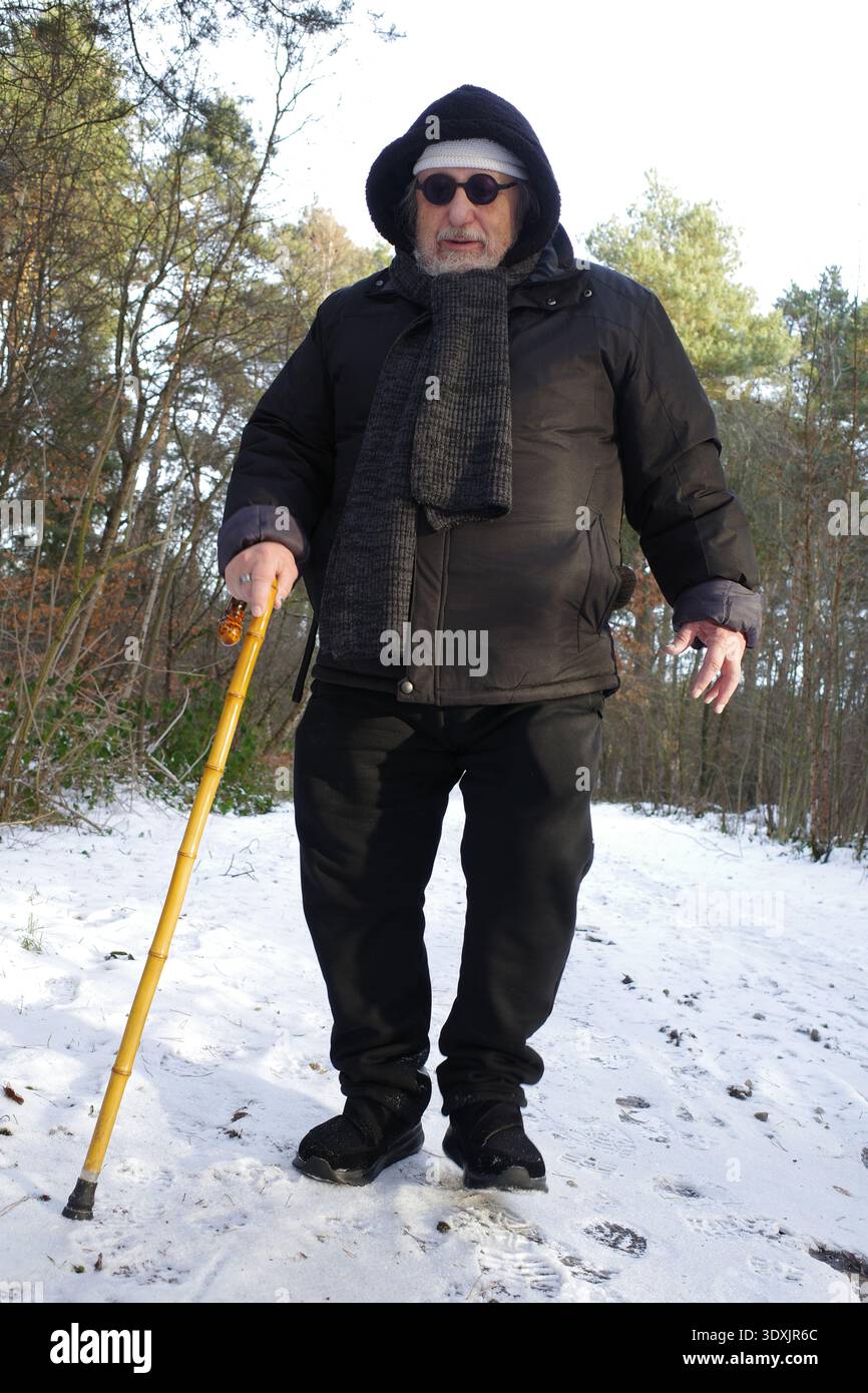 Un homme aîné marche sur un sentier enneigé dans une zone boisée. Il porte un manteau d'hiver noir, une capuche, une écharpe et des lunettes de soleil. Il tient un CA de marche jaune Banque D'Images
