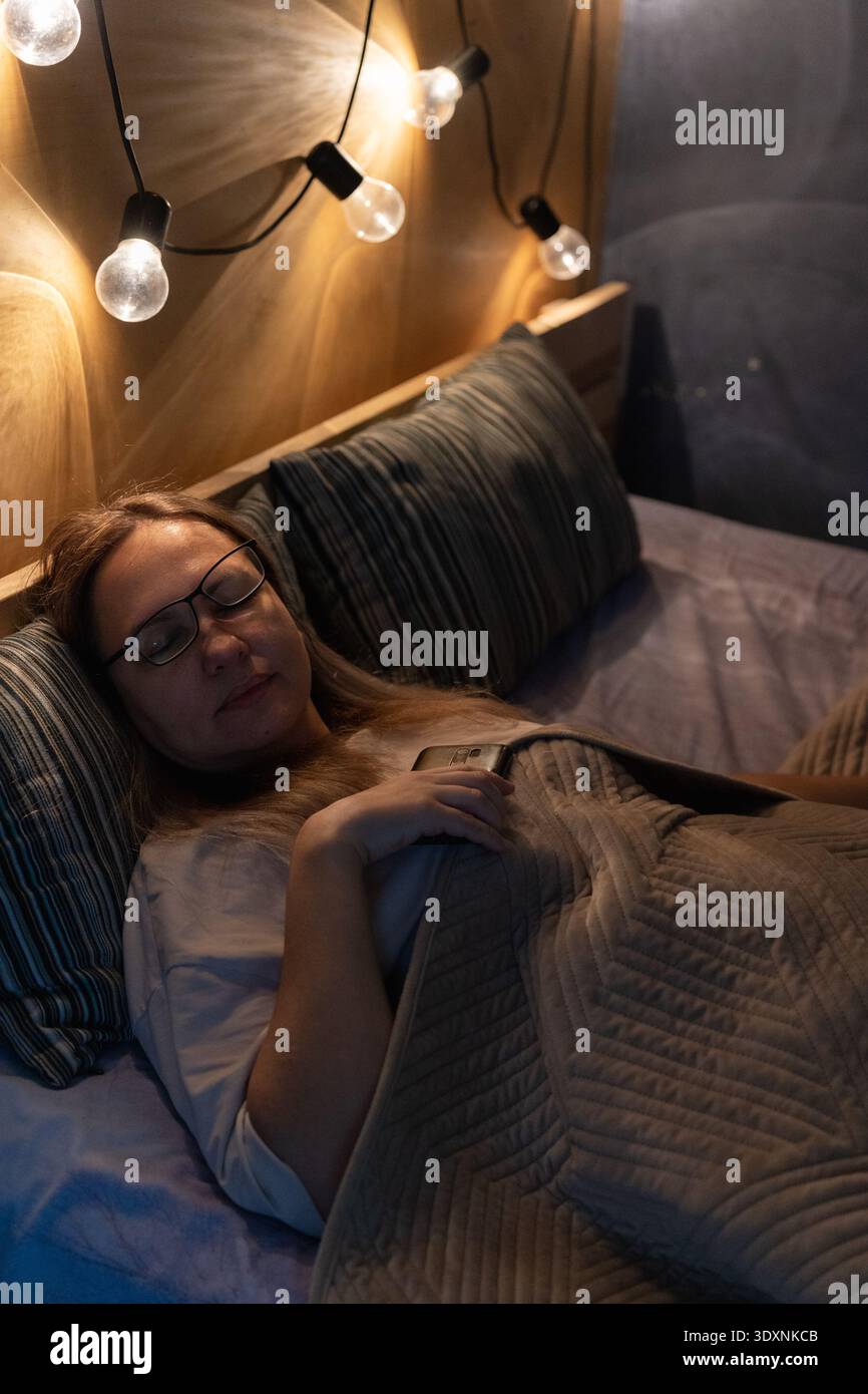 Femme dormant dans son lit la nuit, tenant toujours son smartphone, souffrant de dépendance numérique et d'insomnie Banque D'Images